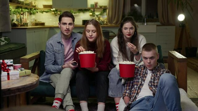 A company of cheerful young men and women relaxes at home, sitting on the couch in the living room. Guy with disability, with amputated hands with friends watching TV and eating popcorn