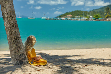 Fototapeta na wymiar A young girl in a bright yellow dress admires the turquoise waters at the beach with palm trees