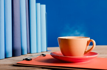 Yellow cup of coffee or tea with hot steam, on note book, with light blue books and blue background.