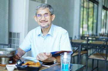 Asian senior man with grey hair sitting in the restaurant with food on table(selective focus at old man face),a smiling traveller read a travel book for guide on his travelling