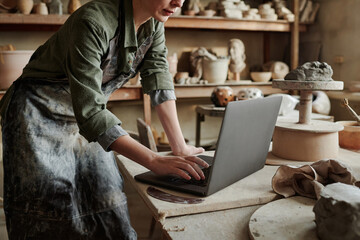 Close-up of craftsperson typing on laptop and creating new models online in the ceramic studio