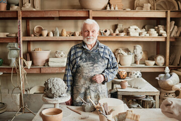 Portrait of senior potter with white beard looking at camera while making sculptures from clay in...