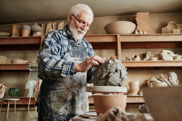 Senior male ceramist working with clay and making dishes by hands in pottery studio - obrazy, fototapety, plakaty