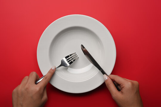 Woman Holding Fork And Knife In Hands With Empty Plate Over Red Background.