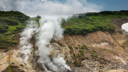 Columns of smoke and steam rise in the thermal valley from fumaroles over the mountain slopes. There are sulfur deposits on the soil, green vegetation on the hill. Blue sky and clouds. Kamchatka.