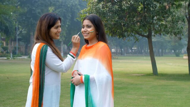 A female artist painting the face of a young girl with the Tricolors of the National Flag. Two ladies / women wearing a white Kurta with Tricolor Dupatta - Republic Day celebrations  patriotism  Na...