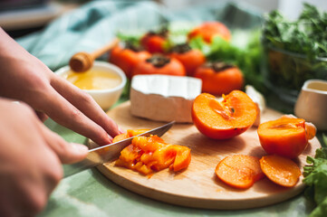 Ingredients and process of making a vitamin winter salad with persimmons, tangerines and cheese Female or teenage hand cuts persimmon on a round wooden board Vegan lunch preparation. Selective focus