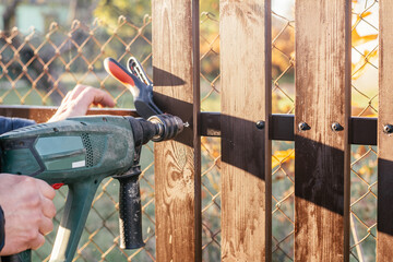 Hand of carpenter holding electric drill for making hole into wooden plank to metal construction for reconstructing wooden fence in sunny day. Outdoor works. Chain-link fence background. Close up.