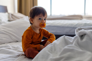 A one-year-old child with a pacifier in his mouth is sitting on the bed in the bedroom.