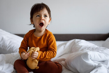 A one-year-old child with a soft toy yawns while sitting on the bed in the bedroom. - obrazy, fototapety, plakaty