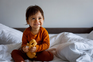 Portrait of a happy little child with a teddy bear sitting on the bed.