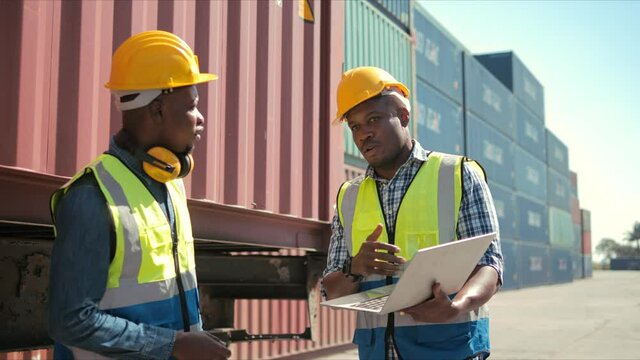 Two professional african engineers wearing safety uniform and hard hats working on laptop in container terminal talking about logistics operations.