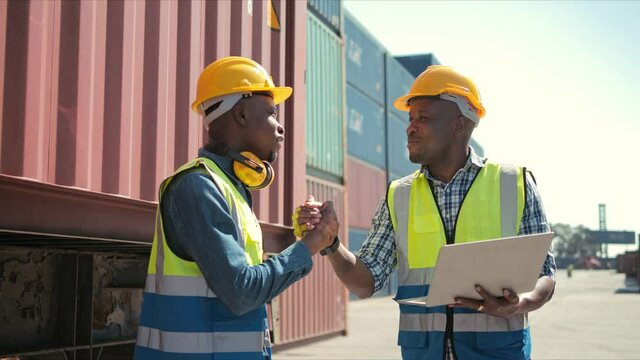 Two professional african engineers wearing safety uniform and hard hats working on laptop and handshake in cargo site industrial worker on factory warehouse background after successful dealing.