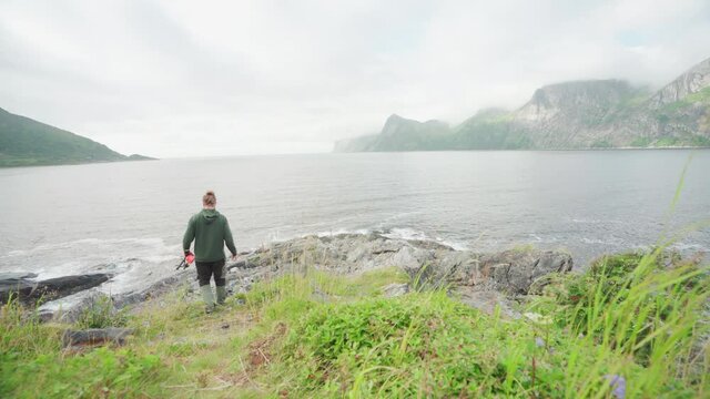 Back View Of A Man Going Fishing Walking Towards A Rocky Shore Holding A Fishing Rod In Segla, Senja Island, Norway. - Wide Shot