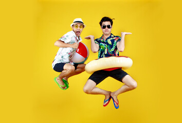 Fototapeta na wymiar Two young Asian guys in summer casual with hat and sunglasses jumping up high with funny gesture with beach ball and ring float