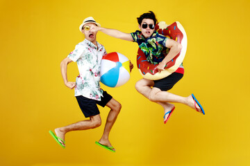 Fototapeta na wymiar Two young Asian guys in summer casual with hat and sunglasses jumping up high with funny gesture with beach ball and ring float