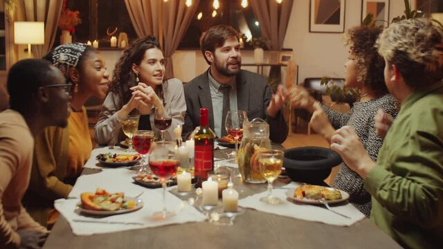 Group of multi-ethnic young friends having fun during birthday party sitting at dining table playing hat game