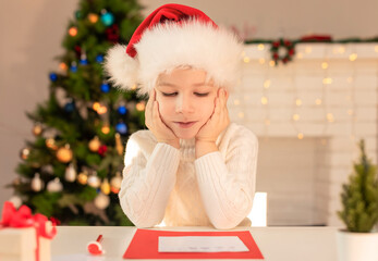 Child boy in red Santa hat writing a letter to Santa Claus. Christmas or New Year cozy holidays concept. Xmas time. Selective focus