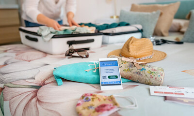 Cell phone with covid passport with unrecognizable woman preparing suitcase for vacation in the background