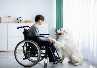 Positive handicapped teenager playing with his dog, holding its paw at home, full length