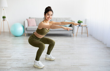 Obraz premium Full length portrait of happy young Indian woman doing squats exercises at home, blank space
