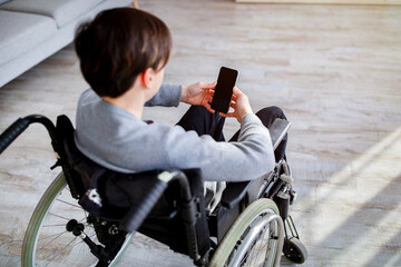 Handicapped teen boy in wheelchair using smartphone with blank screen indoors, mockup for mobile...
