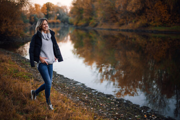 Naklejka na ściany i meble Beautiful young woman walking towards river looking away smiling with teeth with black jacket on shoulders, hands on sweater with trees covered with sunset in background. Autumn walk in forest.