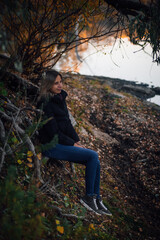 Fototapeta premium Shining young woman sitting on tree roots in forest, smiling, looking away and posing near river with tree in background, wearing blue jeans, black jacket and grey sneakers. Autumn walk in forest.