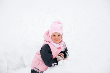 Fototapeta na wymiar Portrait photo of female kid looking into camera and smiling sitting in snowdrift in winter in park wearing warm winter clothes. Astonishing background full of white color and snow. 