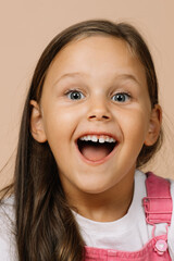 Little girl with shining eyes and excited smile with teeth, opened mouth and raised eyebrows looking at camera wearing bright pink jumpsuit and white t-shirt on beige background.