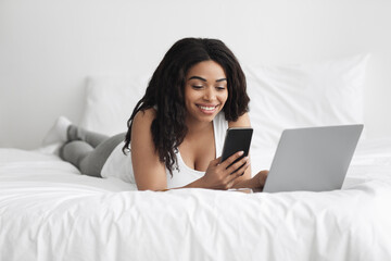 Two factor authentication. Happy black woman using smartphone and laptop computer in bed