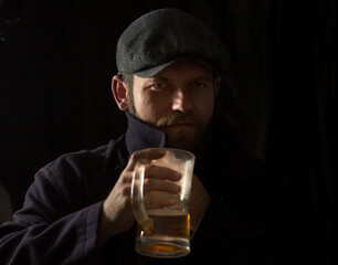 a man with a beard in a vintage cap and coat with the collar turned up drinks beer in the dark from...