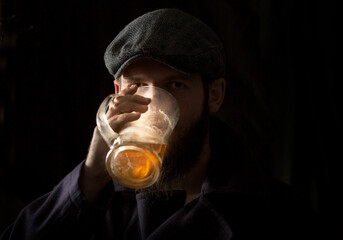 a man with a beard in a vintage cap and coat with the collar turned up drinks beer in the dark from a mug. Front view.