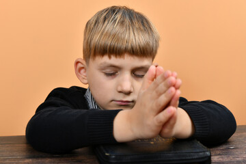 A boy is praying on his knees with folded hands on a closed Bible.