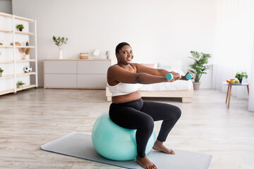 Plus size black woman working out with dumbbells on fitness ball at home, empty space
