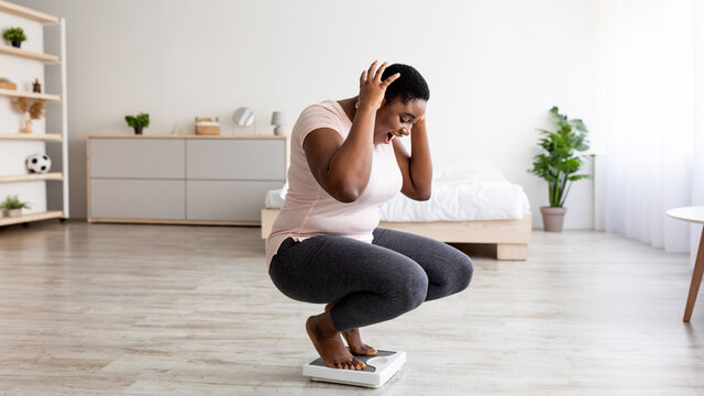 Plus size black woman cannot believe her weight loss result, shouting OMG, sitting on scales at home, feeling excited