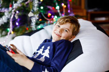 Cute little blond kid boy playing with a video game on gadget console on Christmas with decorated...