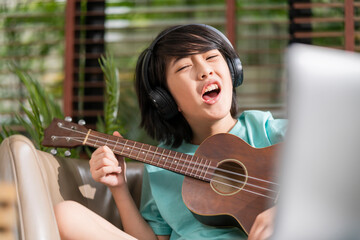 asian child boy sing lound while learn online ukulele study course with fun and exiting at...