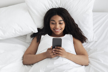Romantic message at morning. Happy young african american lady lying in bed under white blanket and using smartphone