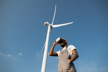 African american man wearing VR headset while standing at rural area with huge windmills around....