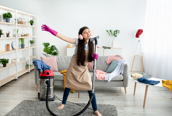 Happy Indian teen girl cleaning her home, wearing headphones, using vacuum cleaner as microphone...