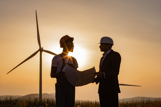 Two multiracial industrial workers examining wind turbines blueprints with amazing sunset on background. Rural area with eco farm of production clean green energy.