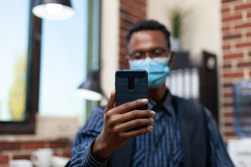 Closeup of startup owner holding smartphone in video call conference wearing covid19 protection mask. Focus on employee smart phone in online interview using wireless earbuds.