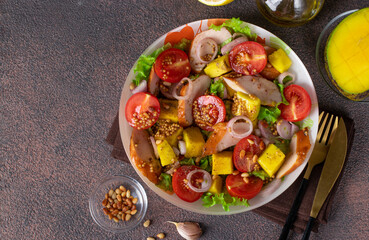 Salad with mango, smoked chicken and cherry tomatoes served with green lettuce leaves in a plate on a brown background, top view