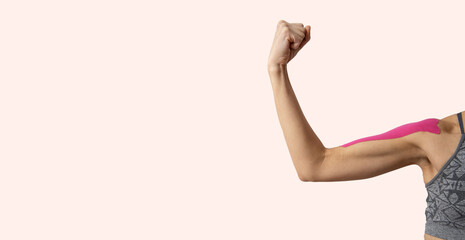 Woman shoulder and pink tape on it.Woman wearing grey sport top and showing kinesio tape on her biceps against bright background - obrazy, fototapety, plakaty