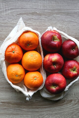 Two sustainable shopping bags filled with apples and oranges on wooden table. Top view.