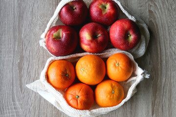 Two sustainable shopping bags filled with apples and oranges on wooden table. Top view.