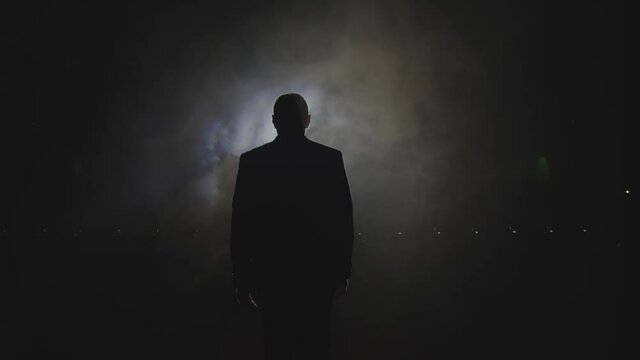 Silhouette of a man in a suit leaves the stage 4K. Mystical atmosphere, smoke, spotlight. Person in the dark
