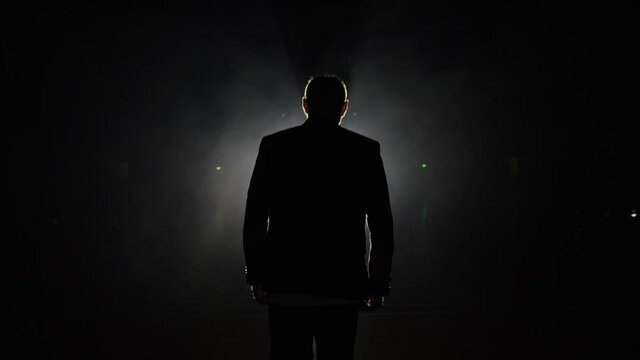 Silhouette of a man in a suit leaves the stage 4K. Mystical atmosphere, smoke, spotlight. Person in the dark