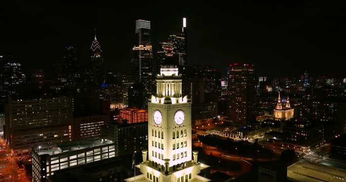 Well lit white clock tower skyscraper among urban city skyline at night. Cinematic establishing aerial shot in darkness downtown city in USA.